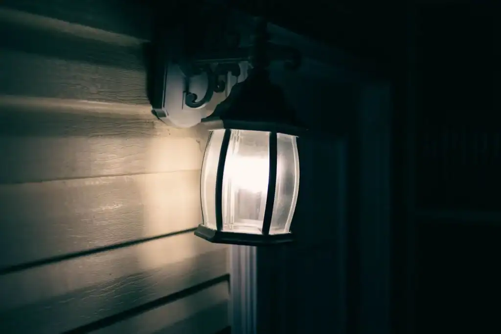 A close-up of a black outdoor wall lantern with clear glass panels and a bright bare bulb glowing against dark siding at night.