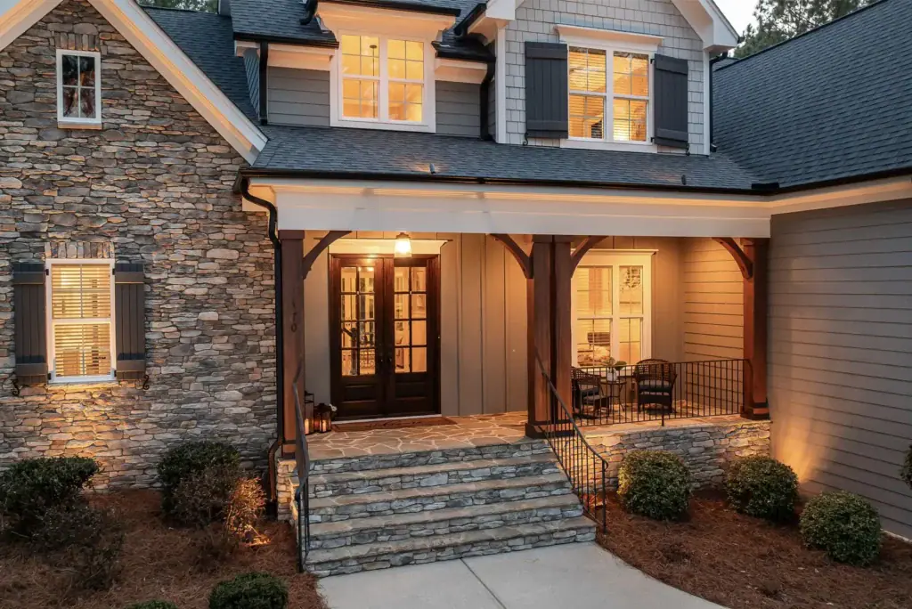 A stone and siding house at dusk with warm lights glowing on the covered front porch above the double front doors.